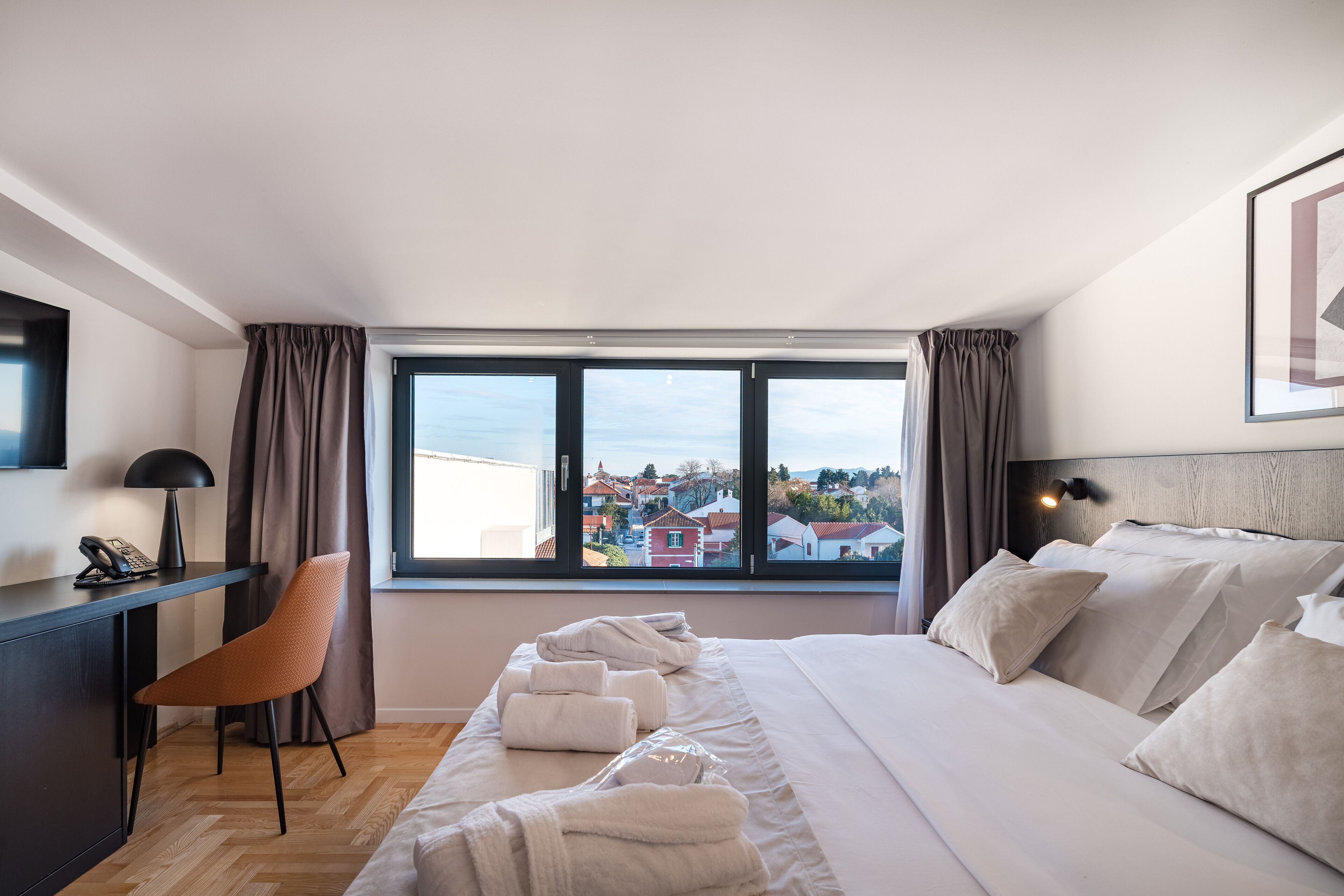 A modern hotel room with a large bed, a desk with a lamp, a chair, and a view of a town through the window.