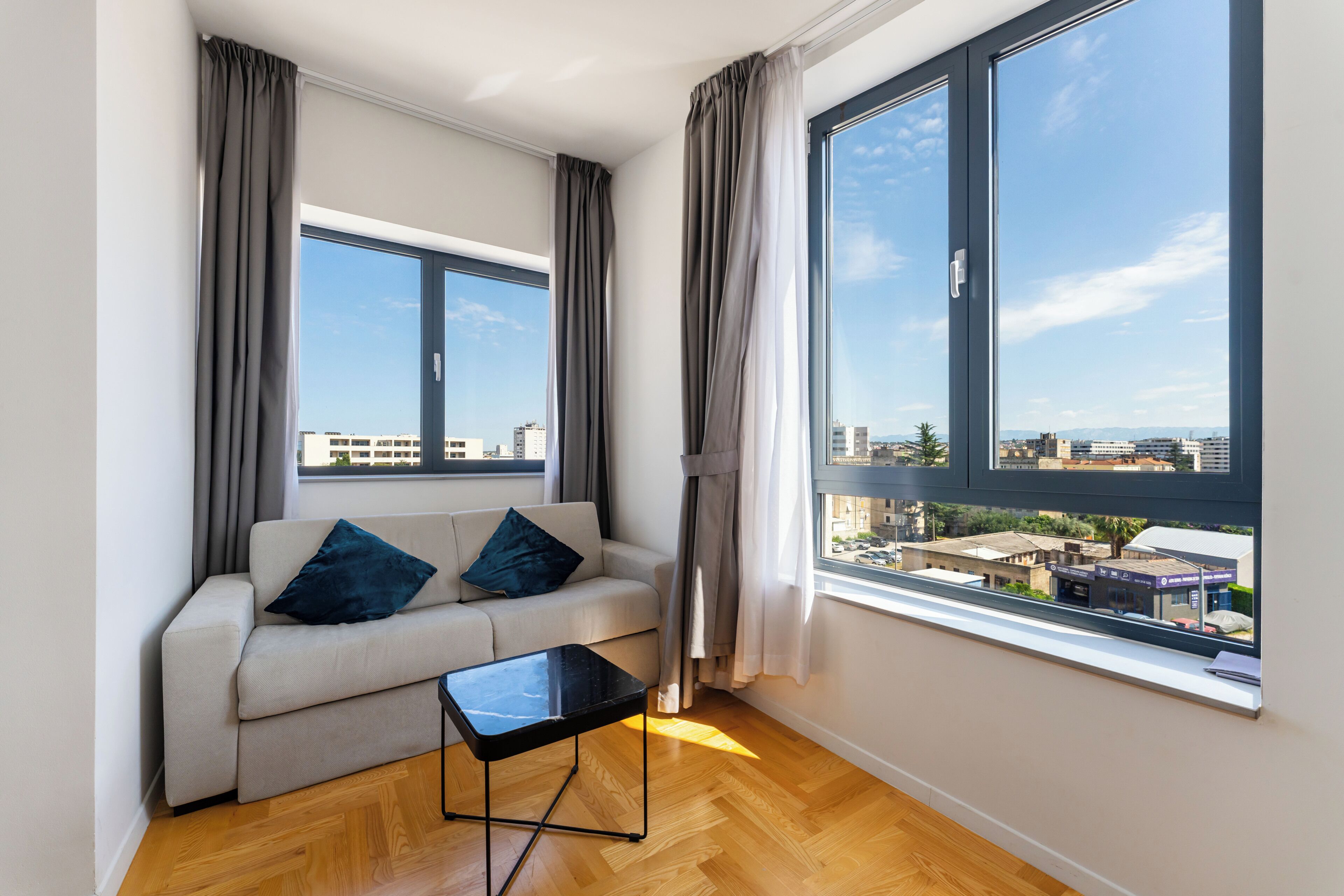 A modern living room with a sofa, a small table, and large windows offering a city view.