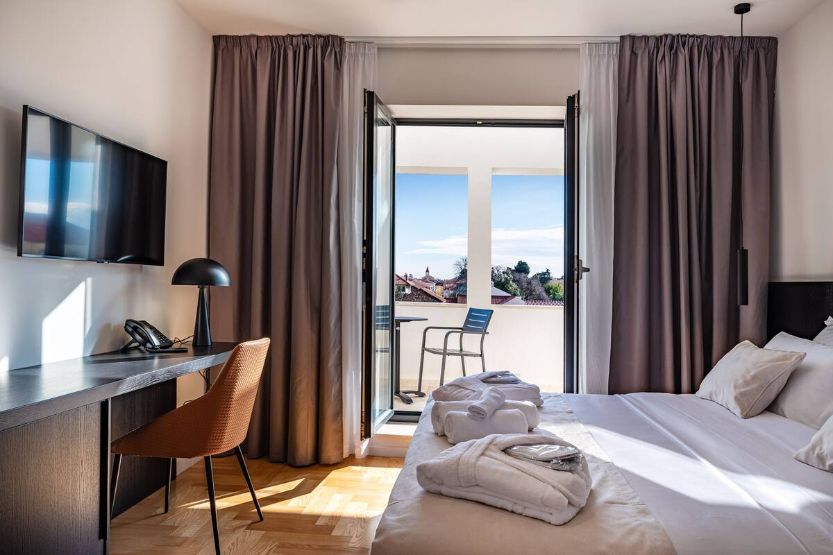 A hotel room with a bed, a desk with a phone, a chair, and a view of the outside.