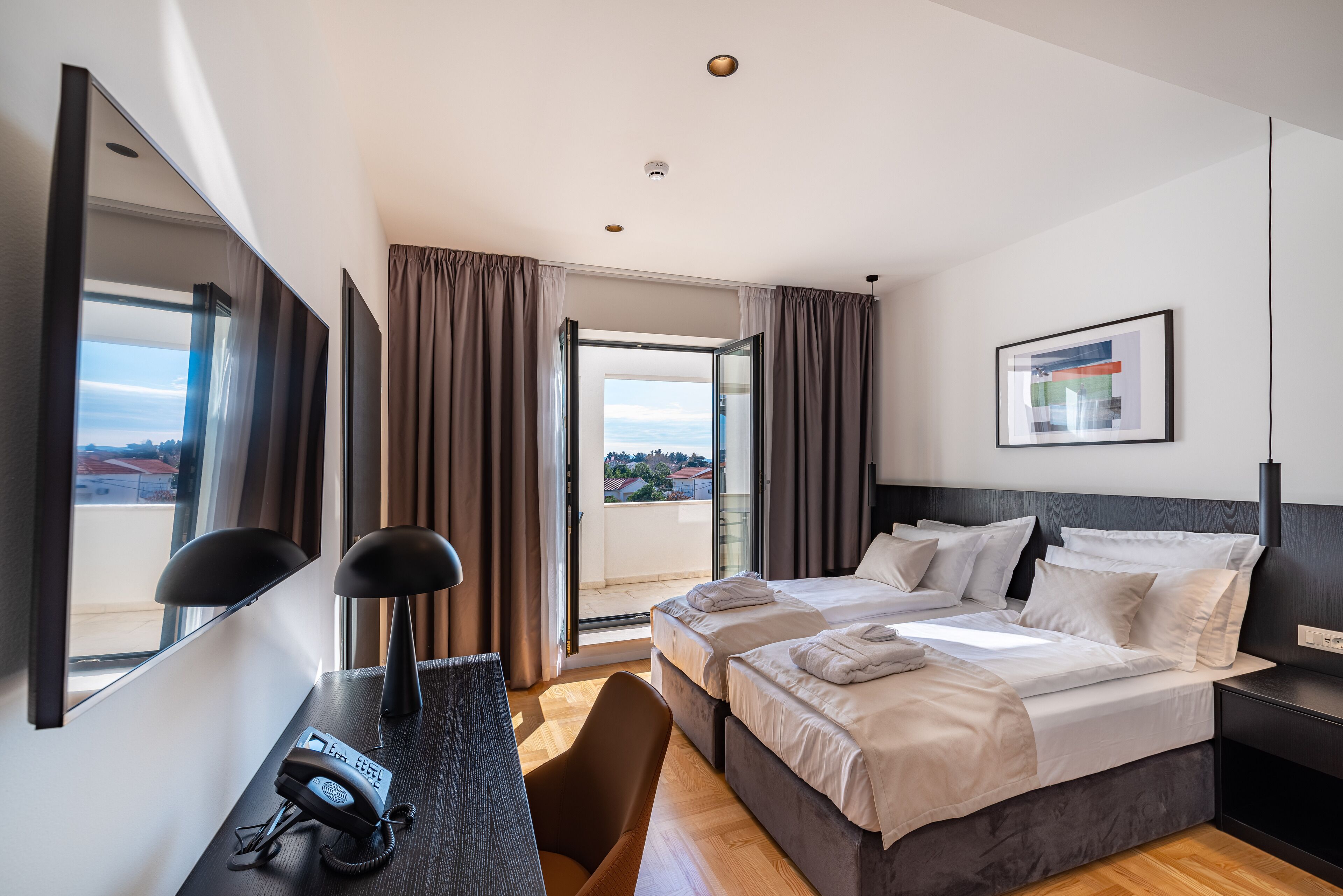 A hotel room with two beds, a desk with a telephone, a chair, a large window, and a painting on the wall.