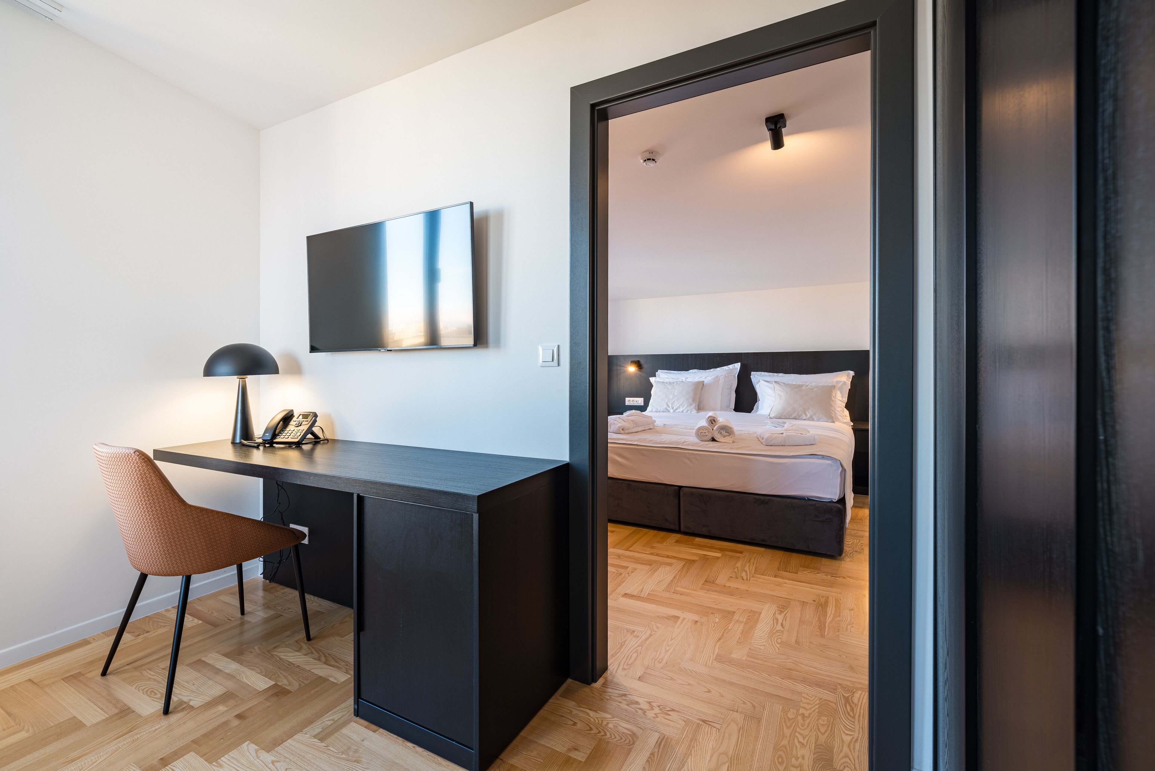 Een moderne hotelkamer met een houten vloer, een bureau met stoel, een bed met wit beddengoed en een grote spiegel.