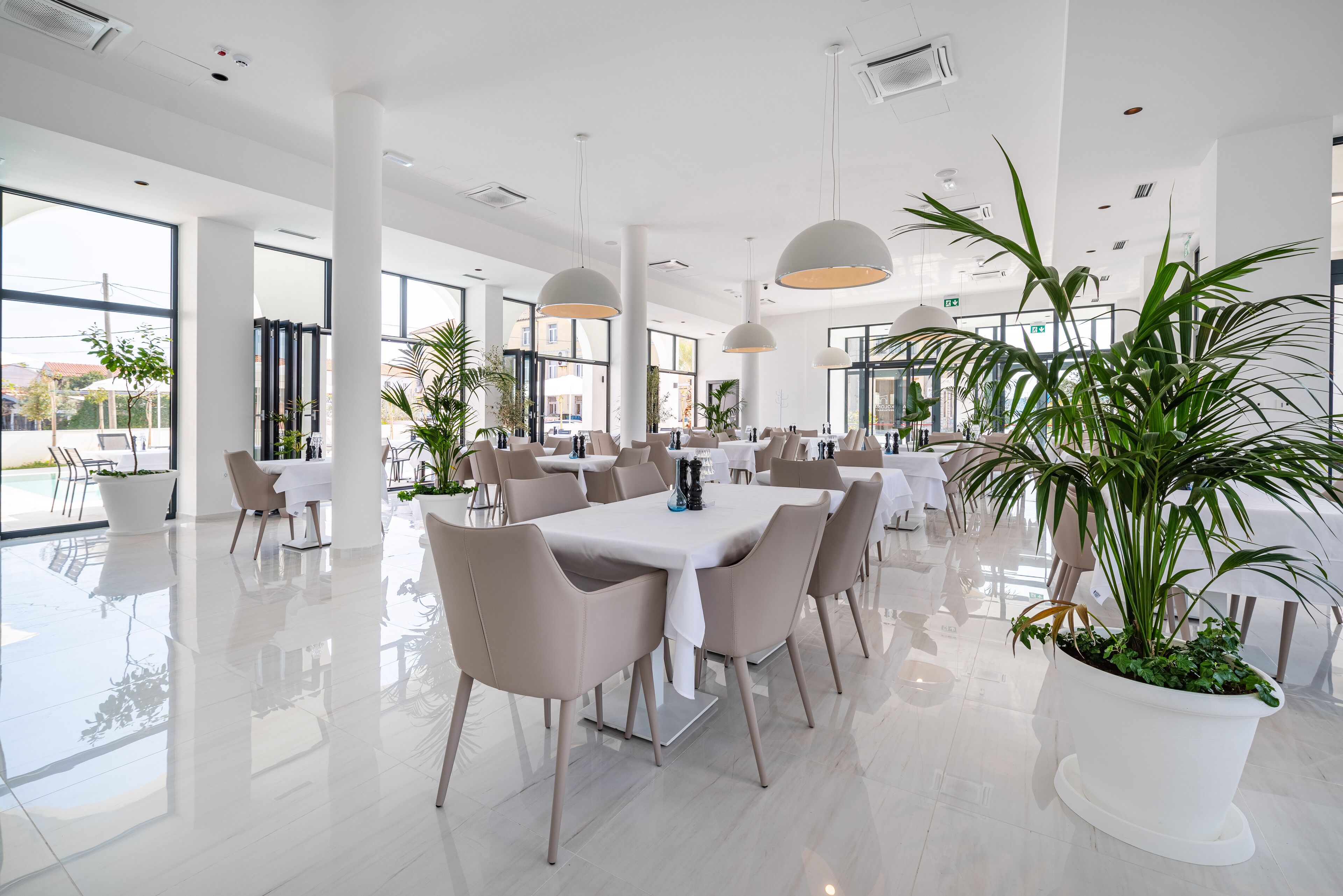 A spacious, modern dining area with white tables and chairs, large windows, and potted plants.