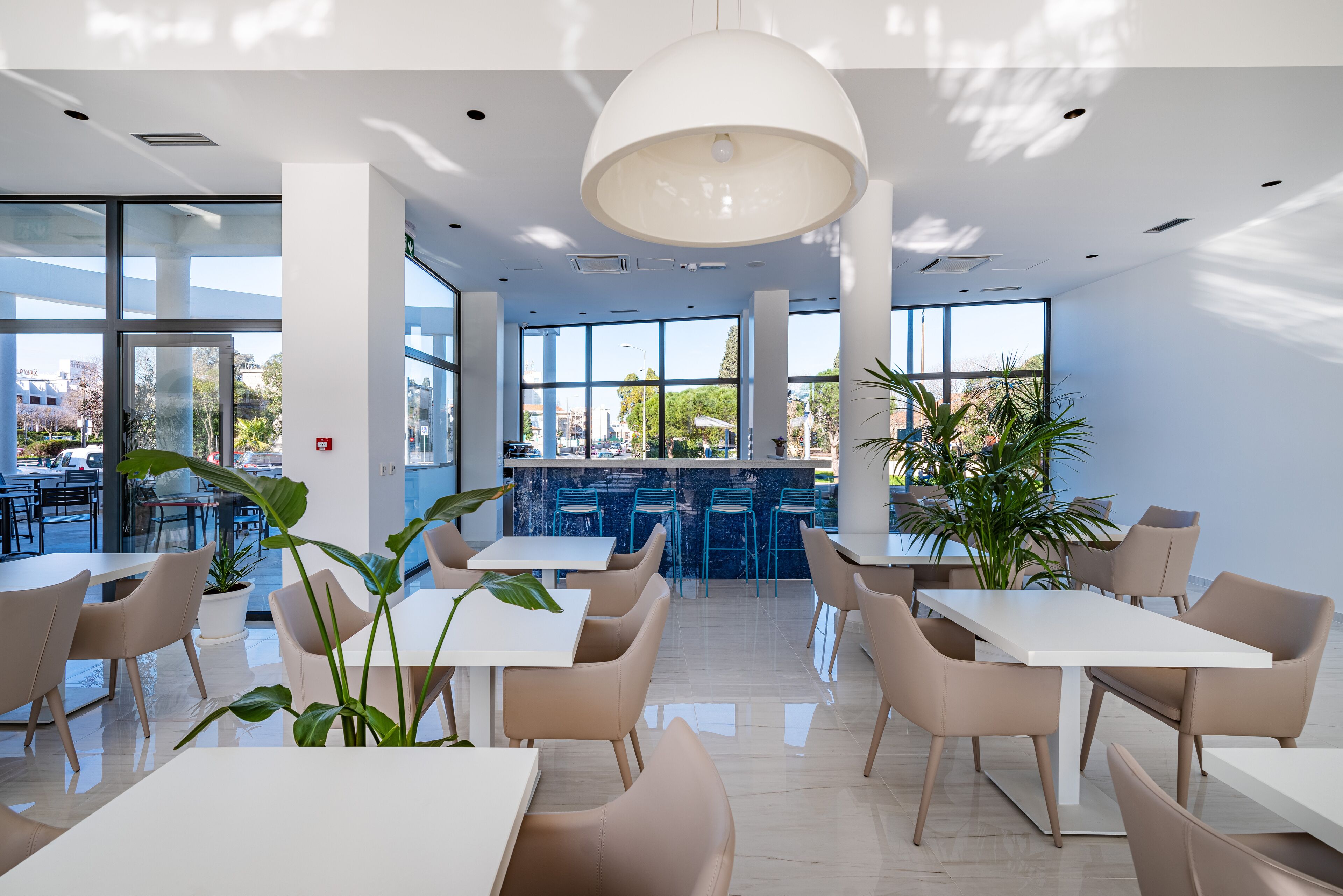A modern indoor dining area with white tables and chairs, large windows, and potted plants.