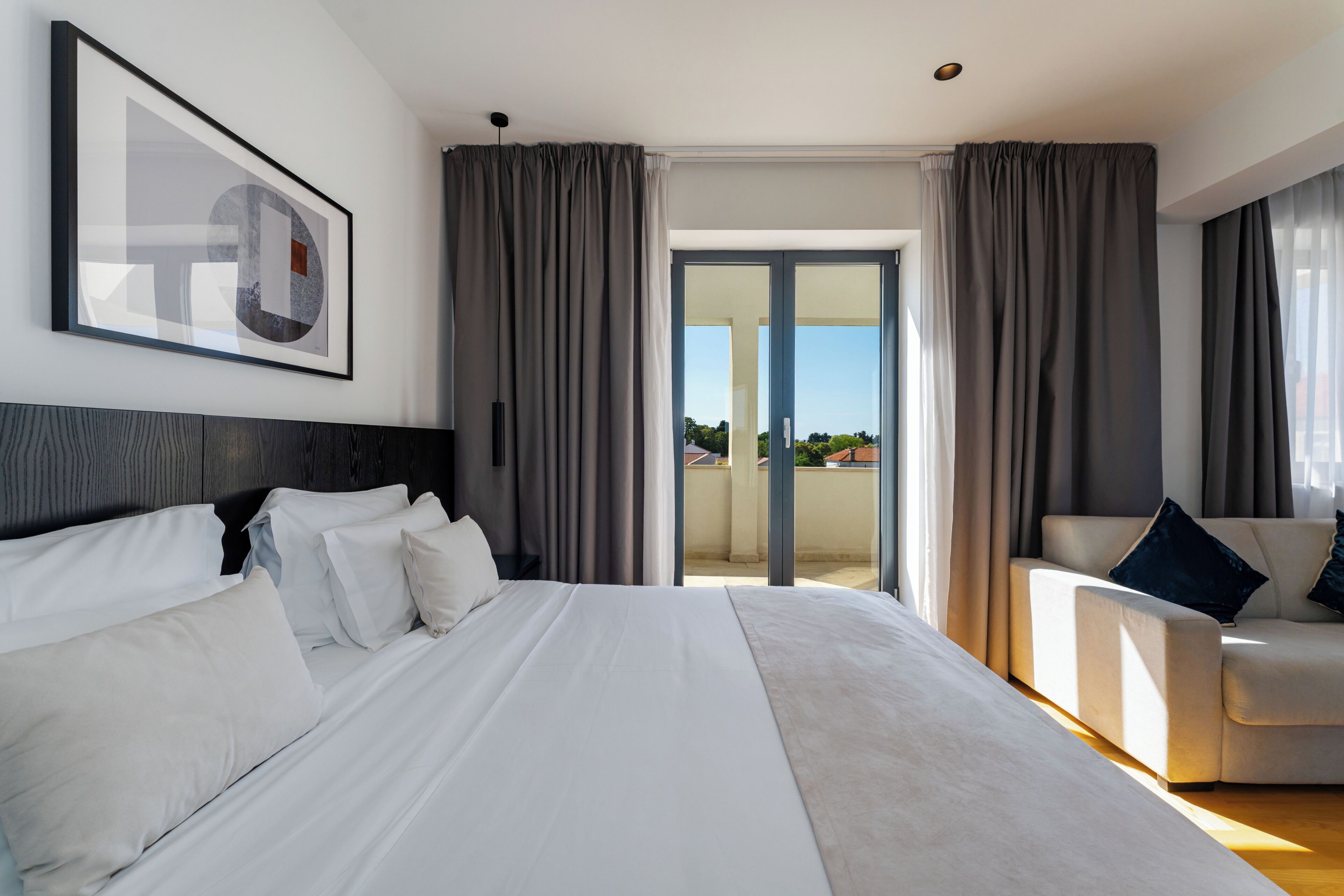 Quarto de hotel moderno com uma cama grande, um sofá e uma varanda com cortinas.