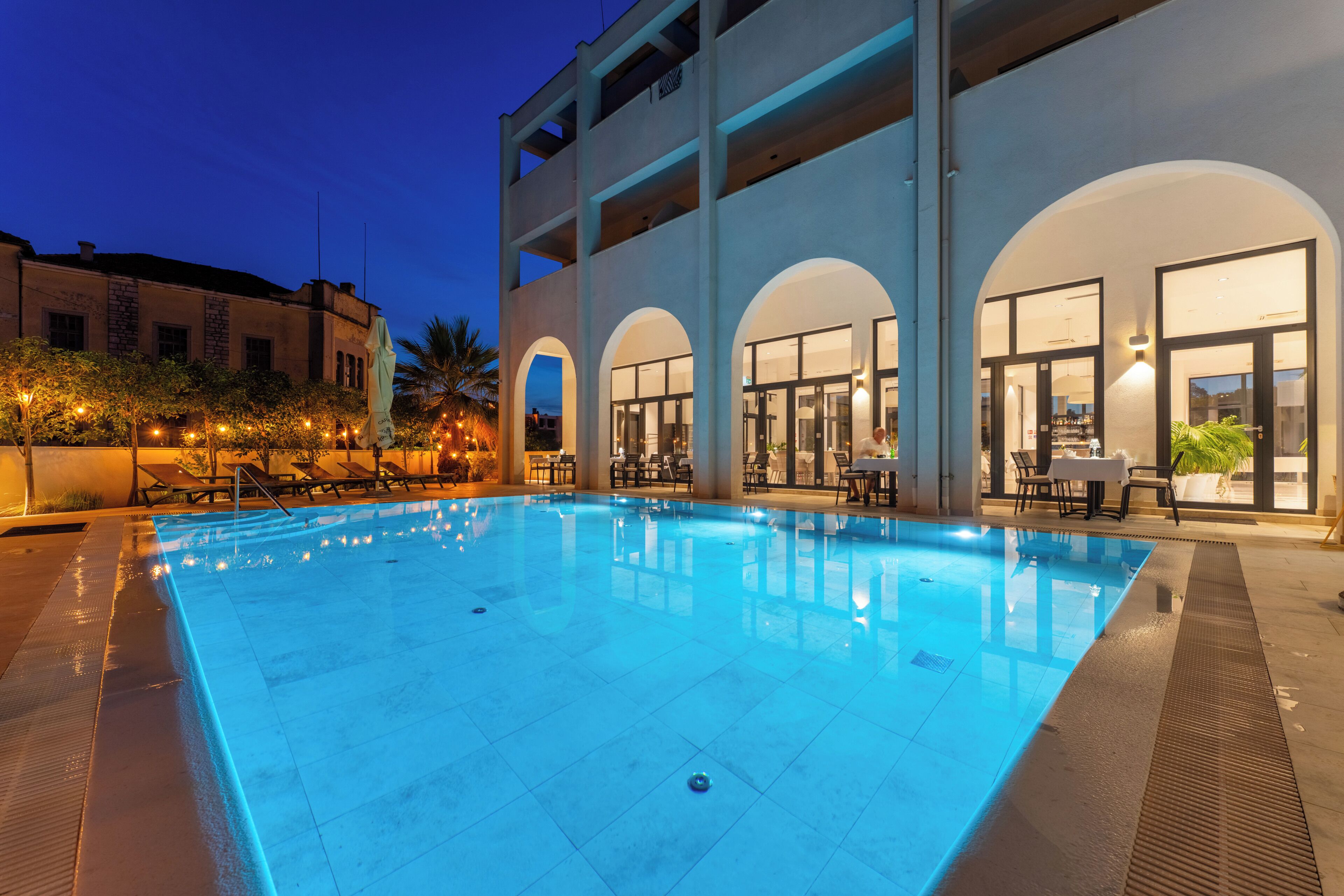 An indoor swimming pool with a clear blue water surface, surrounded by lounge chairs and tables, and a building with multiple balconies and large windows.