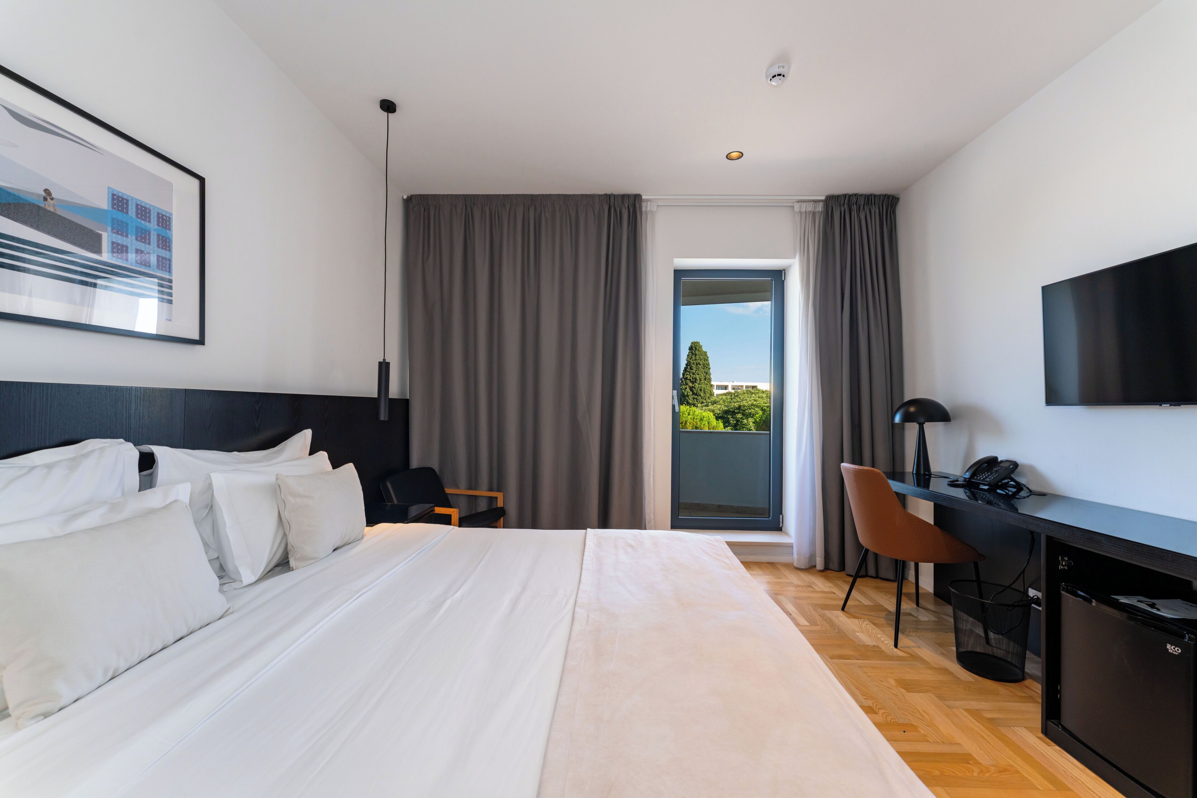 A hotel room with a bed, a desk, a chair, a TV, and a view of greenery outside.