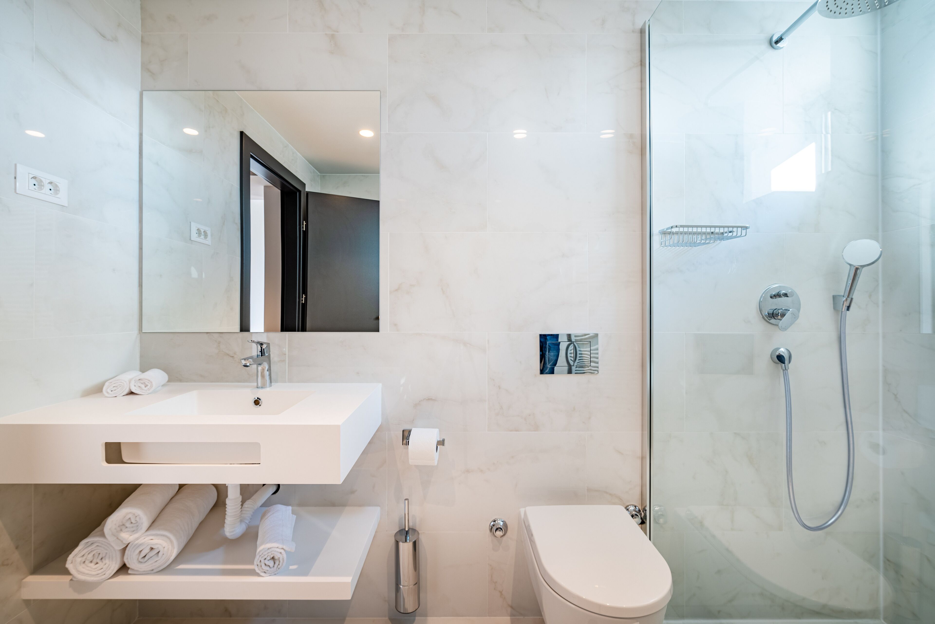 Un bany modern amb un lavabo, un mirall i un vàter blancs. Hi ha una dutxa amb porta de vidre i una barra de tovalloles amb tovalloles.