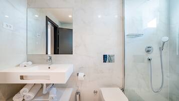Un baño moderno con lavamanos, espejo y váter blancos. Cuenta con una ducha con puerta de vidrio y un perchero con toallas.