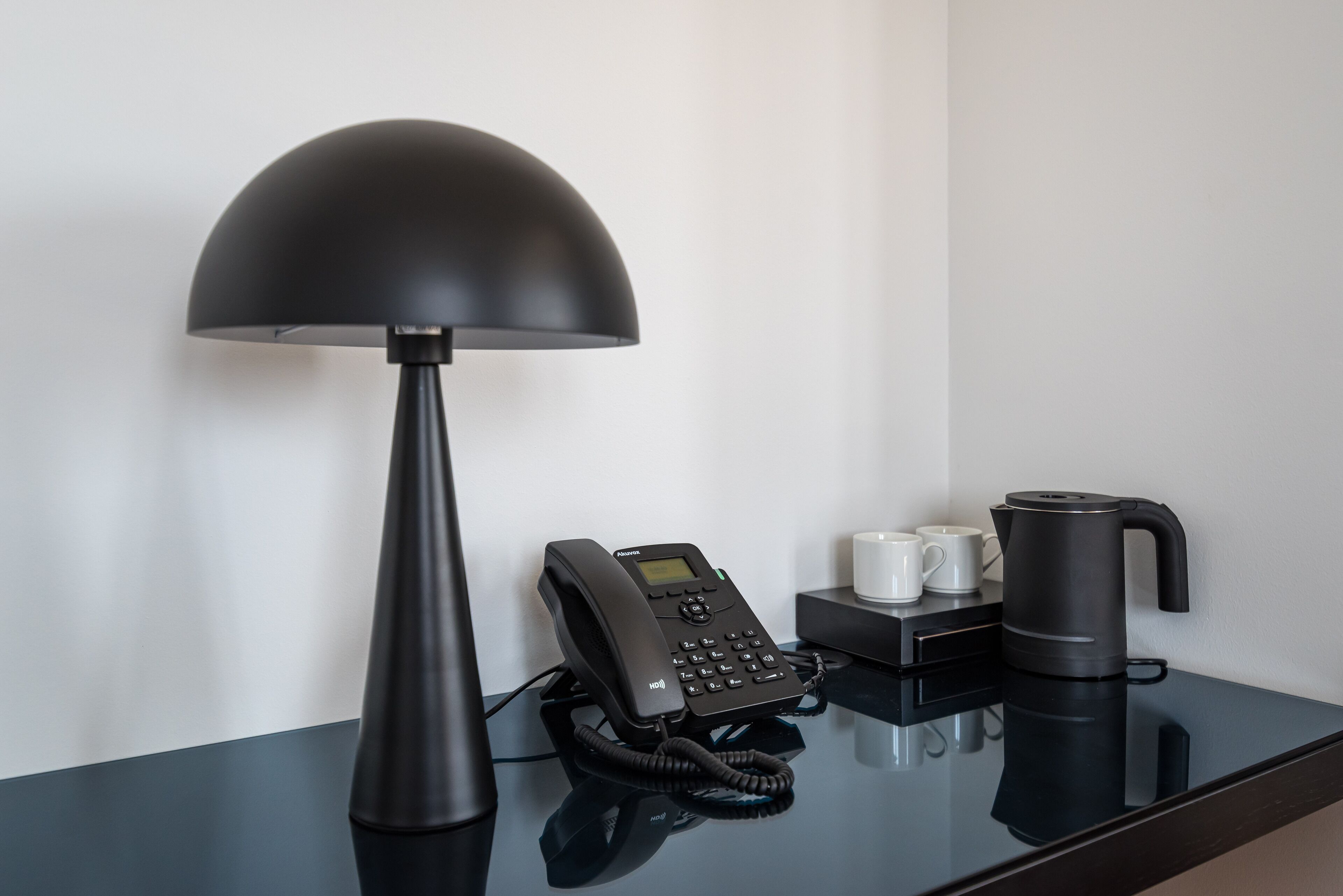Et moderne hotelværelse med et sort skrivebord, en skrivebordslampe, en telefon, to hvide kopper og en kaffemaskine.