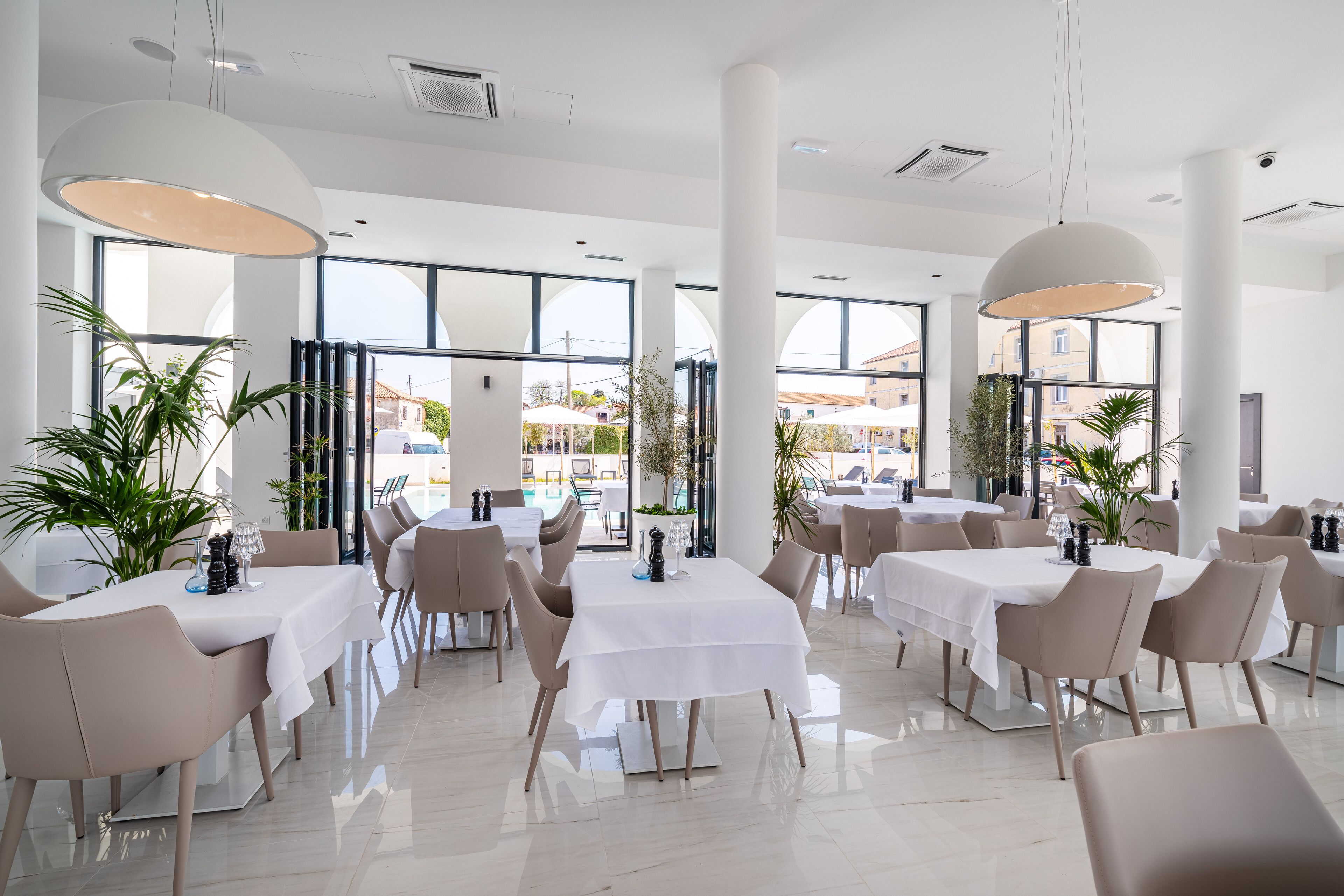 A modern dining area with white tables and chairs, large windows, and indoor plants.