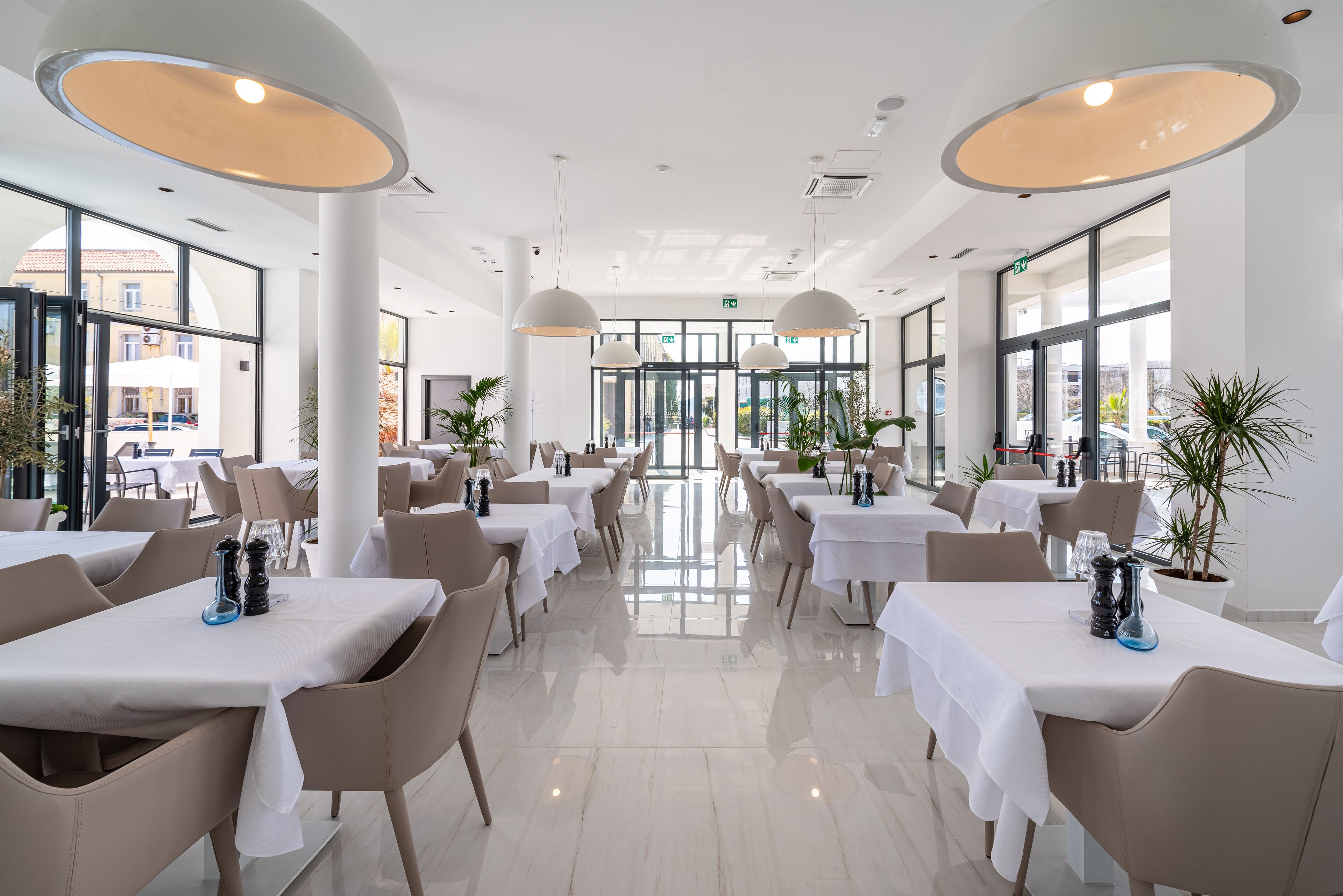 Un ristorante moderno con tavoli e sedie bianche, ampie finestre e luci a sospensione.