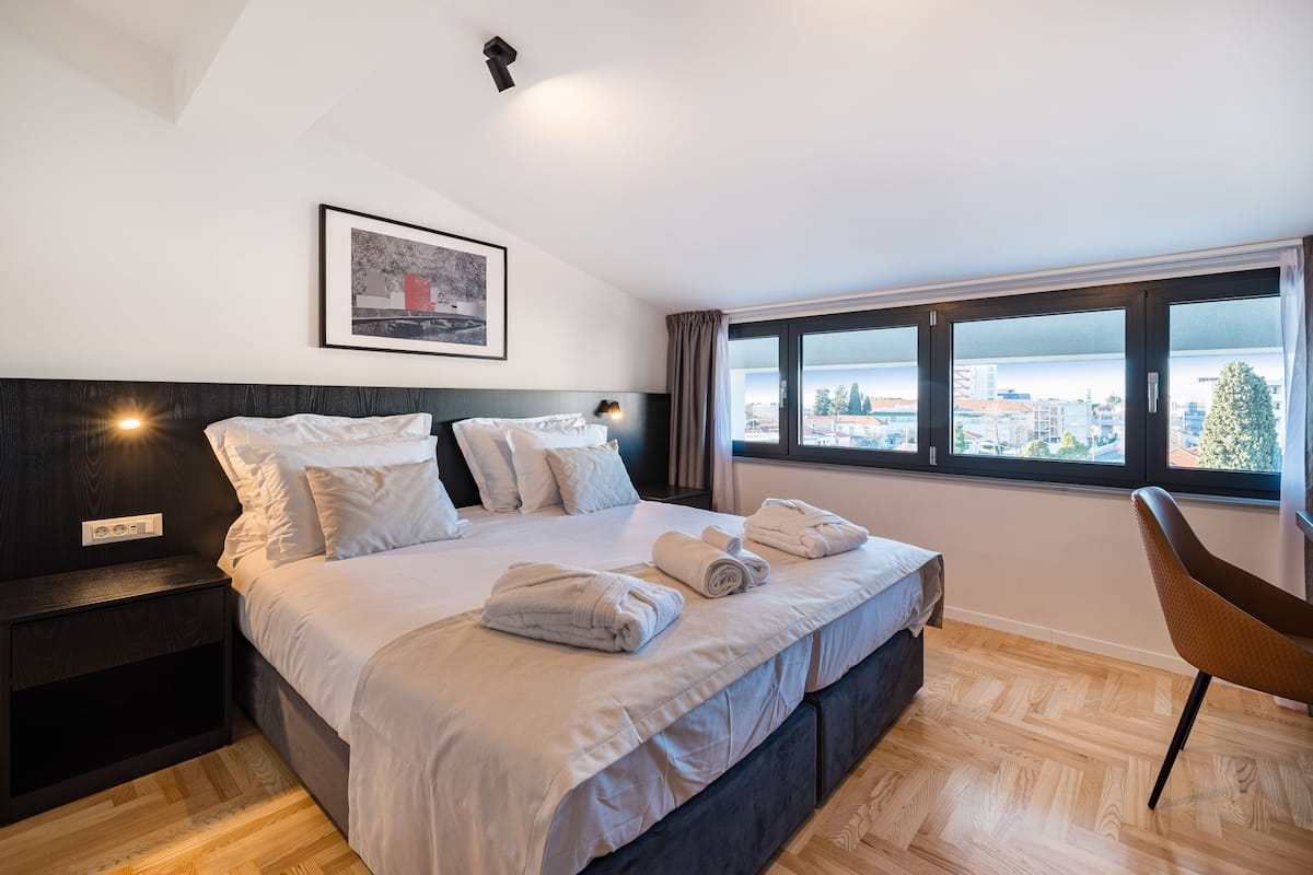 A modern hotel room with a large bed, a nightstand, a chair, and a window with a city view.