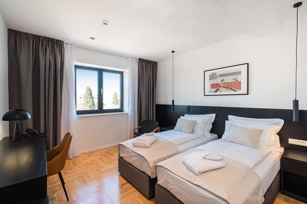 A hotel room with two beds, a desk, a chair, a window with curtains, and a framed picture on the wall.