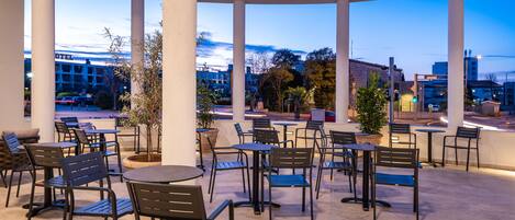 An outdoor patio area with multiple tables and chairs, surrounded by white columns, and overlooking a cityscape.