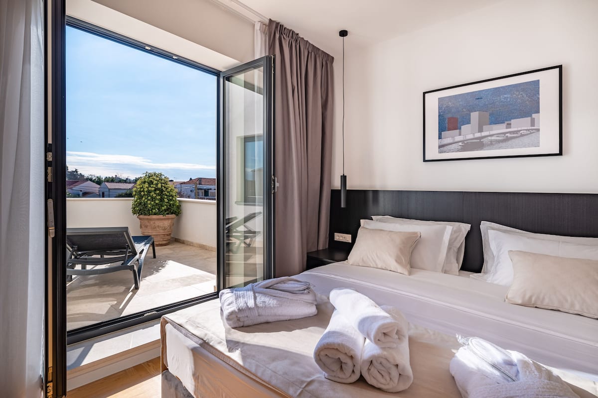 A hotel room with a bed, a large window, a potted plant, and a framed picture on the wall.