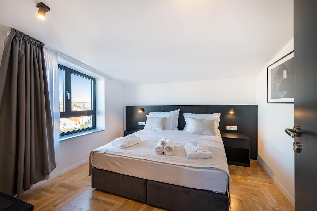 A modern hotel room with a large bed, a nightstand, a window with curtains, and a city view.