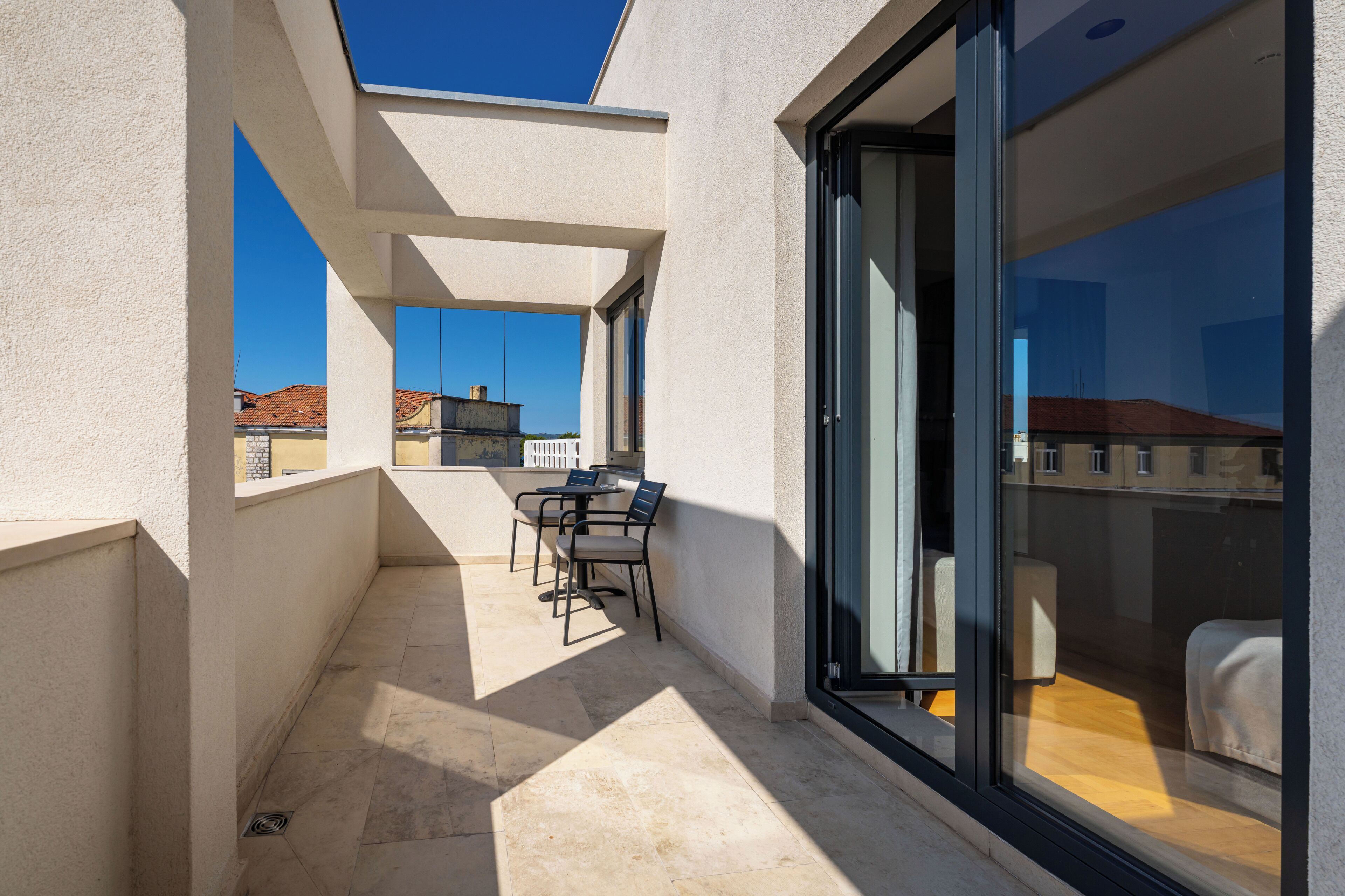 A sunlit balcony with a table and chairs, a sliding glass door, and a view of buildings in the background.