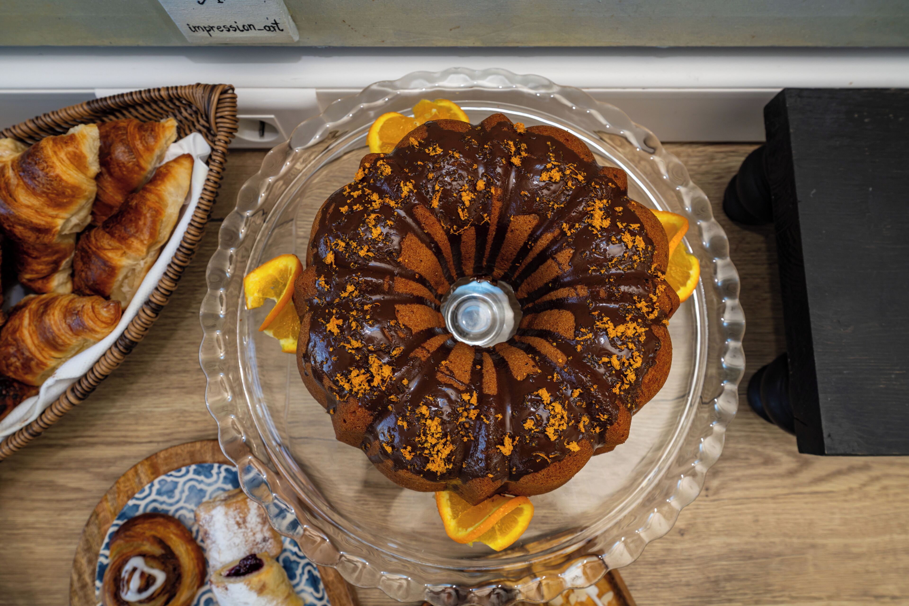 Un pastel bundt sobre un expositor de vidrio, con rodajas de naranja, rodeado de pastelería sobre una mesa de madera.