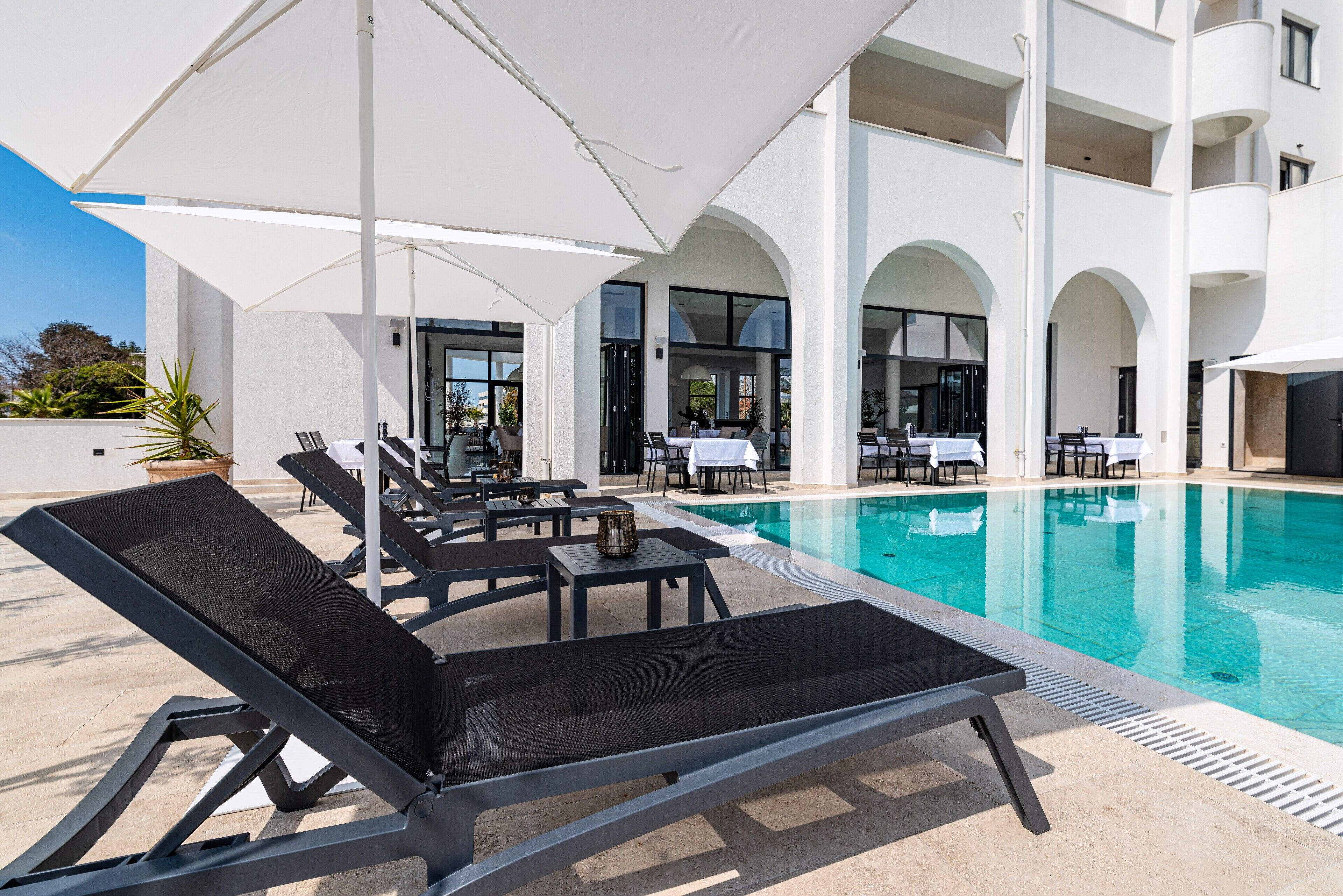 Un área junto a la piscina con sillas de descanso y sombrillas, una piscina y un edificio con ventanas arqueadas y balcones.