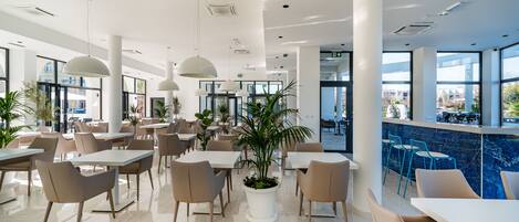 Un espace intérieur vaste et lumineux, avec de nombreuses tables et chaises, de grandes fenêtres et des plantes en pot.