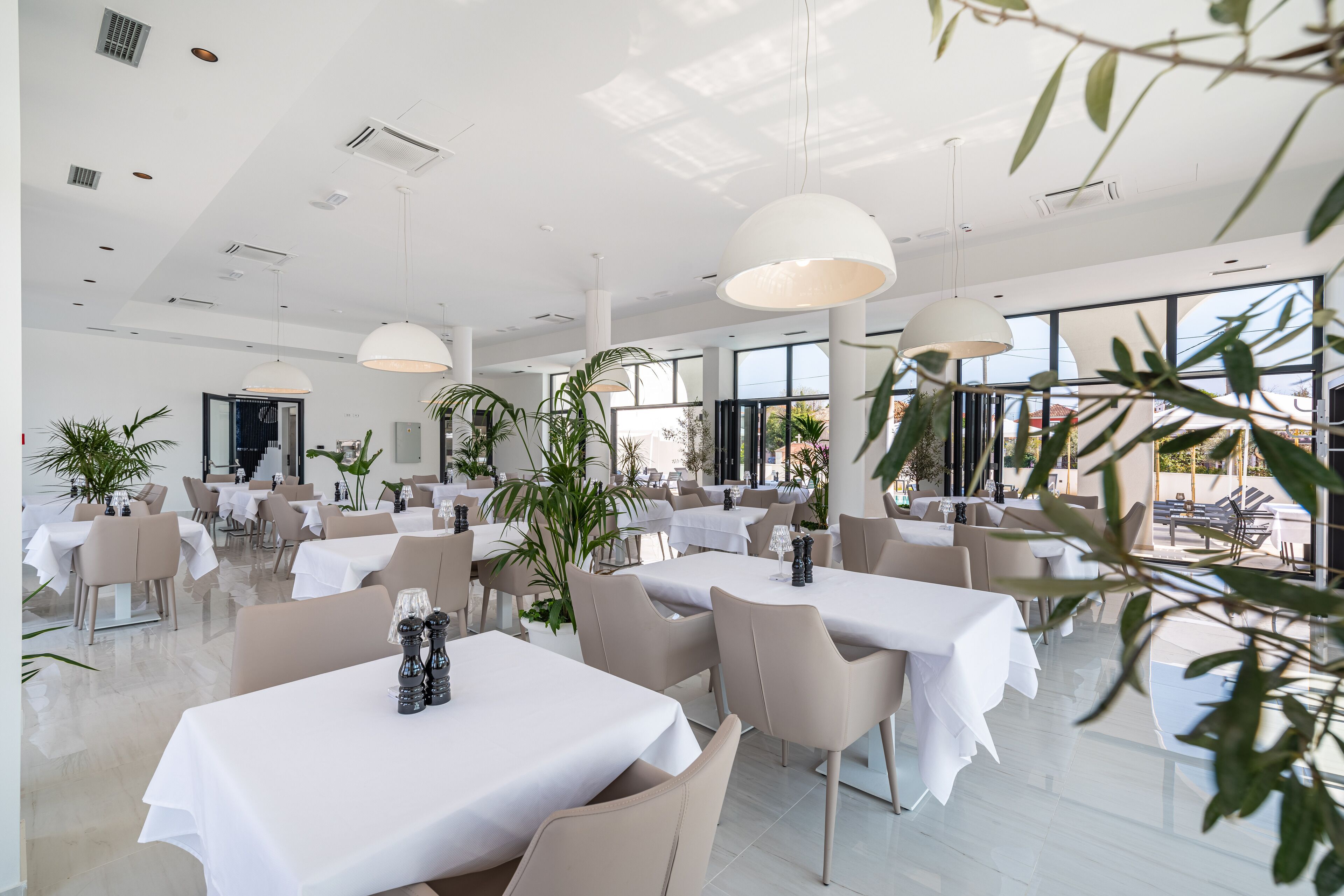 A spacious dining area with multiple tables and chairs, large windows, and indoor plants.