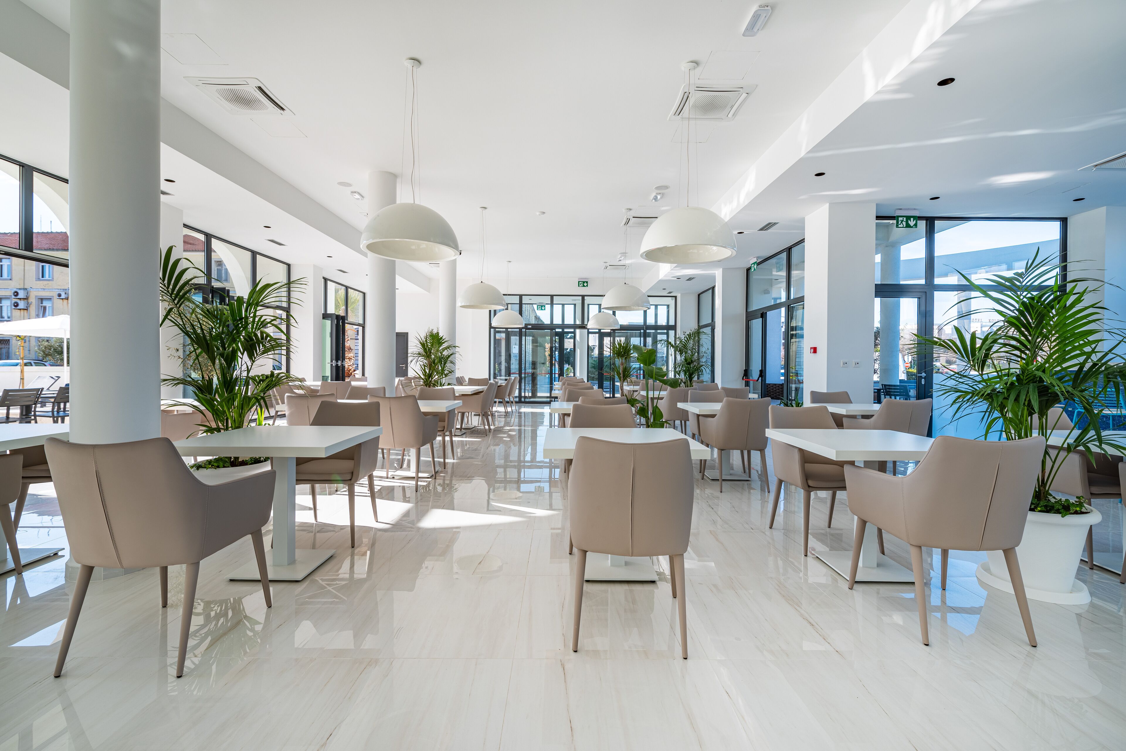 A spacious indoor area with multiple tables and chairs, large windows, and potted plants.