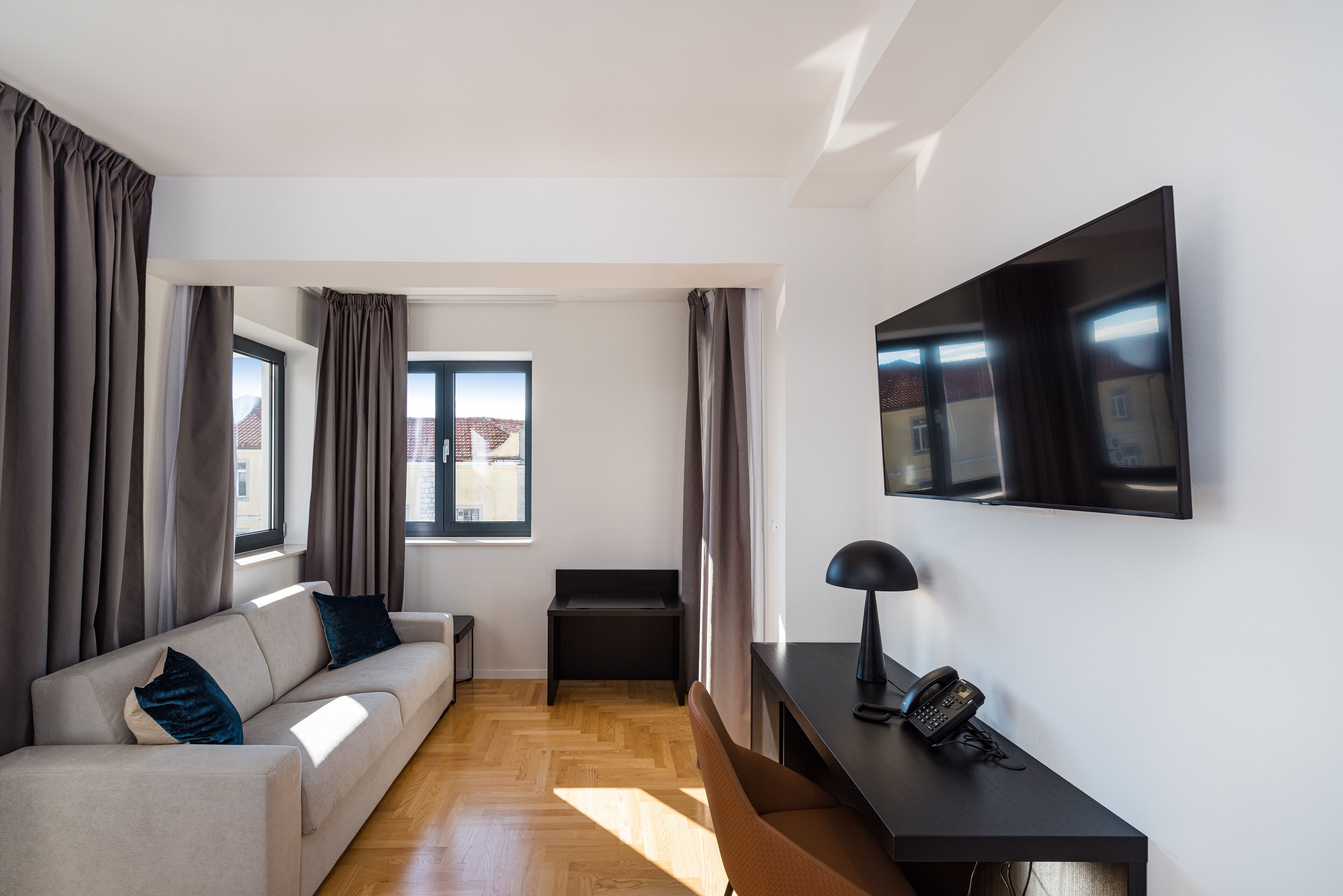 Ein modernes Wohnzimmer mit einer Couch, einem kleinen Tisch mit Lampe, einem Telefon und einem an der Wand befestigten Fernseher.