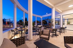 An outdoor patio area with multiple tables and chairs, large white columns, and a view of the cityscape at dusk.