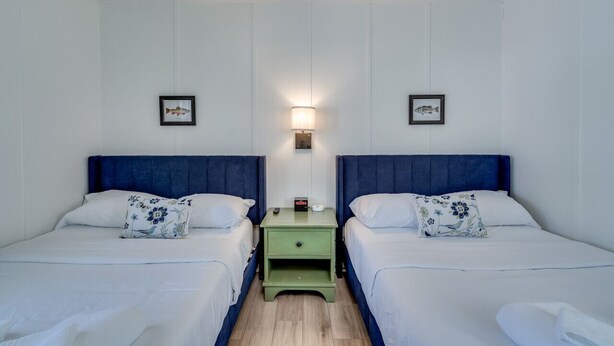 Ein Doppelzimmer mit zwei Betten, beide mit blauen Kopfteilen, weißer Bettwäsche und blumigen Kissen. Zwischen den Betten steht ein grüner Nachttisch mit Lampe und Uhr.