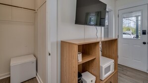 Una habitación con un armario blanco, un televisor de pantalla plana montado, una estantería de madera con un microondas y una puerta blanca con ventanal.