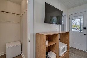 Une pièce avec une armoire blanche, un téléviseur à écran plat fixé au mur, une étagère en bois avec un four à micro-ondes, et une porte blanche avec une fenêtre.