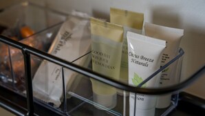 Una repisa de vidrio con artículos de aseo, incluyendo un tubo etiquetado 'Citrus Breeze Naturals' y una bolsita etiquetada 'Good Day Bonjour Fleurs Dais'.