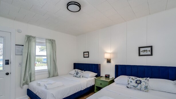 Una habitación con dos camas, una ventana con cortinas, una lámpara de pared y cuadros enmarcados.