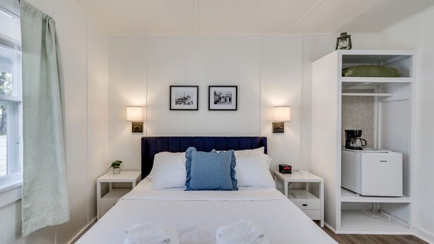 Une chambre d’hôtel avec un lit, des tables de chevet, une petite table d’appoint, une lampe, une petite étagère avec une machine à café et un tableau encadré au mur.