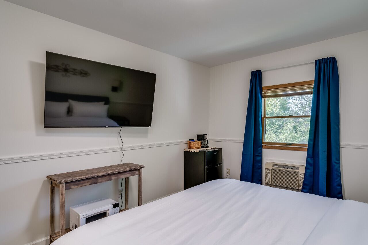 Ein Hotelzimmer mit einem Bett, einem flachbildfernseher an der Wand, einem kleinen Tisch mit einer Mikrowelle, einem schwarzen Schrank und einem Fenster mit blauen Vorhängen.