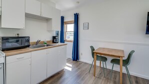 A compact kitchen with white cabinets, a microwave, a coffee maker, and a sink. There is a wooden table with two chairs and a window with blue curtains. - K's Motel with Stunning St. Lawrence River Views - Room 9 (Clayton)