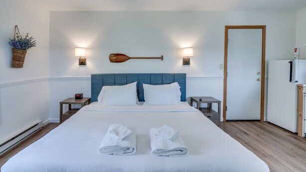 A neatly made bed with two towels, a wooden paddle wall decoration, two wall-mounted lamps, a small bedside table with a clock, and a door to the right. - K's Motel with Stunning St. Lawrence River Views - Room 9 (Clayton)