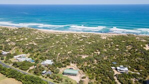 Vista aérea de una zona costera con vegetación densa, una playa y varios edificios, incluyendo una casa grande con techo verde y una estructura más pequeña con techo azul.