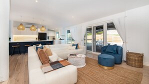 Una sala de estar moderna con un sofá, una mesa de centro y un sillón azul. Hay una zona de cocina con armarios blancos y una zona de comedor con una mesa y sillas.