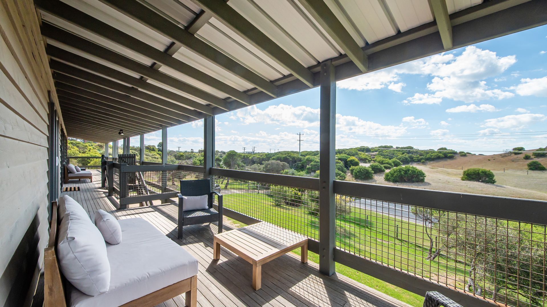 A covered balcony with a wooden deck, a sofa, a chair, and a view of a green landscape with trees and a road.