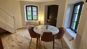 A dining area with a round table and chairs, a window with a view of greenery, and a door in the background. - Le Parc du Séquoia : stay in a former apiary (Ostheim)