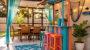 An outdoor patio with a wooden table and chairs, a colorful bar area, and a potted plant.