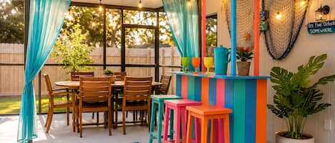 En udendørs terrasse med et træbord og stole, et farverigt barområde og en potteplante.