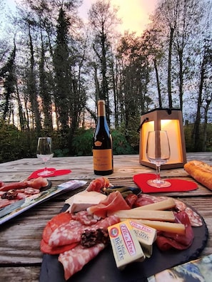 Ein Picknick mit einer Weinflasche, Gläsern sowie verschiedenen Käsesorten und Fleischwaren auf einem Holztisch in einem Wald.