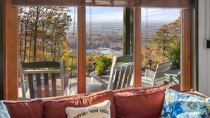 Habitación con un amplio ventanal que ofrece una vista panorámica de un bosque y un lago. Hay dos sillones de mimbre y un sofá con un cojín que dice 'SUPPORT YOUR LOCAL'.
