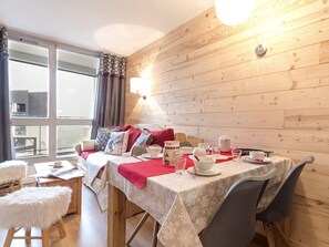 Quarto aconchegante com parede de madeira, cama com roupa de cama vermelha, mesa de jantar posta com pratos e xícaras, e um banquinho branco fofo.
