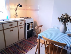 A compact kitchen with white cabinets, a gas stove, an oven, a striped rug, and a table with a blue checkered tablecloth.