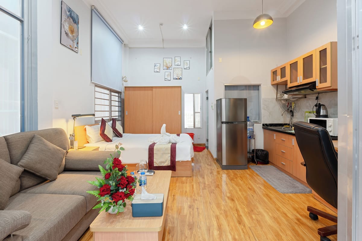 Ein modernes Hotelzimmer mit einer Couch, einem Bett, einem kleinen Tisch mit Blumenvase und einer Küchenzeile mit Holzschränken und einem Kühlschrank.