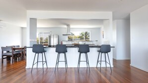 A modern kitchen with a central island, bar stools, and a dining area with wooden chairs. - Omaroo - Absolute waterfront location (ILUKA)