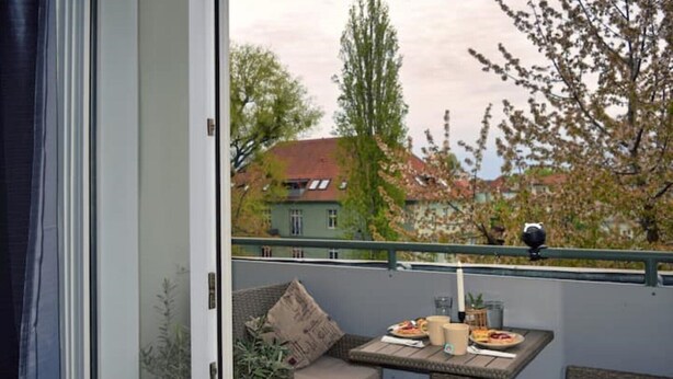 Un balcon agrémenté de meubles en osier, une table avec des assiettes et des tasses, et une vue sur des immeubles et des arbres.